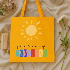 Peut inclure: Sac fourre-tout jaune avec un motif de soleil et le texte "you are my sunshine". Le sac a un design simple avec une couleur jaune ensoleill&eacute;e et des poign&eacute;es assorties. Le texte est dans une police color&eacute;e et ludique.