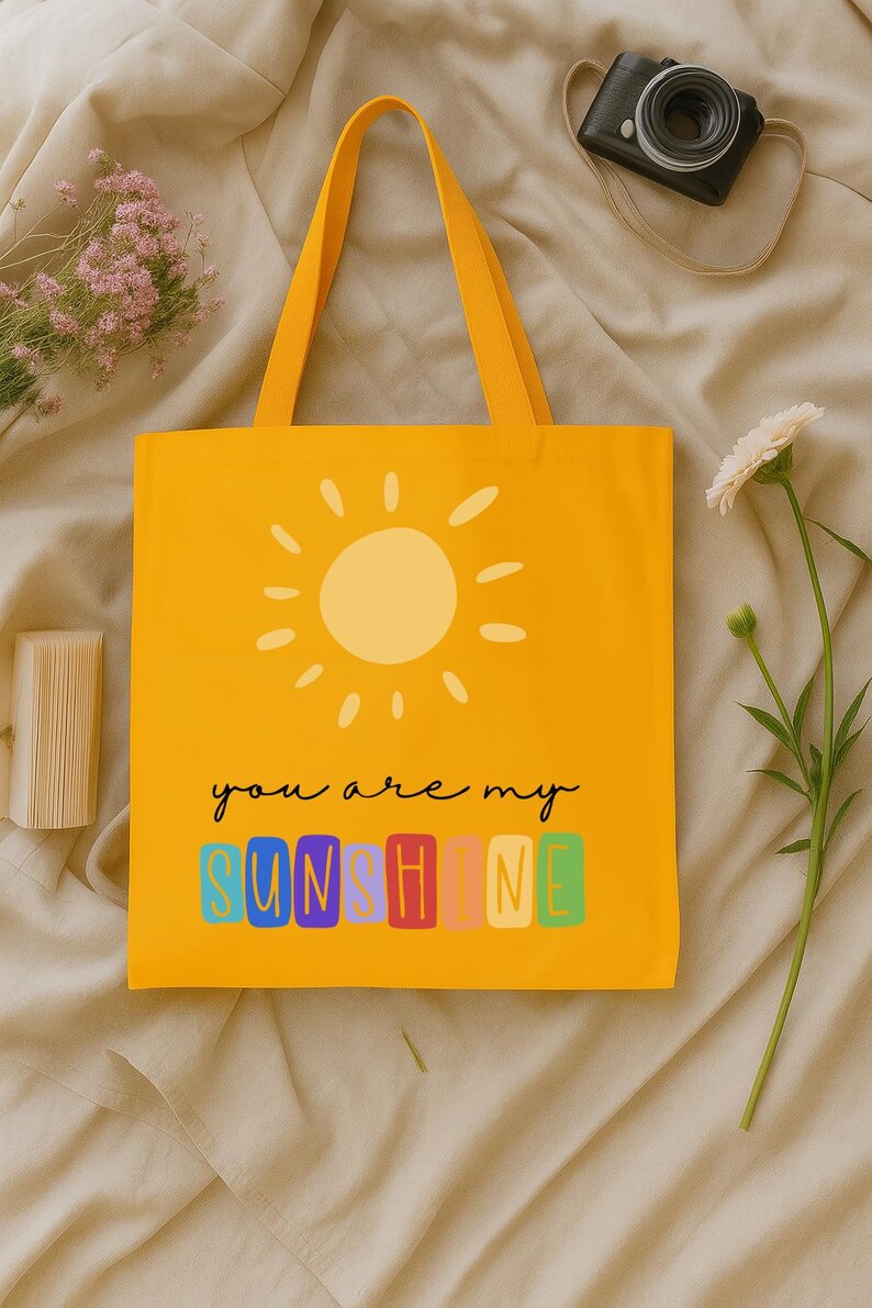 Peut inclure: Sac fourre-tout jaune avec un motif de soleil et le texte "you are my sunshine". Le sac a un design simple avec une couleur jaune ensoleill&eacute;e et des poign&eacute;es assorties. Le texte est dans une police color&eacute;e et ludique.