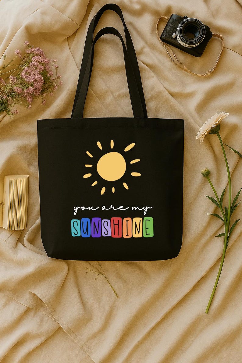 Peut inclure: Sac fourre-tout noir avec un soleil jaune et l'inscription "you are my SUNSHINE" en blanc et en couleurs arc-en-ciel. Le sac a des anses noires et est fait d'un mat&eacute;riau r&eacute;sistant.