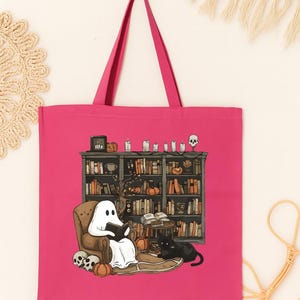 Puede incluir: Un bolso tote rosa intenso con una ilustraci&oacute;n de Halloween. Un fantasma lee en un sill&oacute;n frente a una estanter&iacute;a, con un gato negro cerca. Calabazas, velas y calaveras se suman a la escena espeluznante.