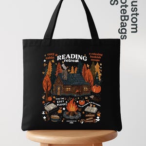 May include: Black tote bag featuring a cabin scene with a campfire and books. Text includes "Reading Retreat", "Cozy Cabin", and "For the Cozy Readers". The design is in autumnal colors.