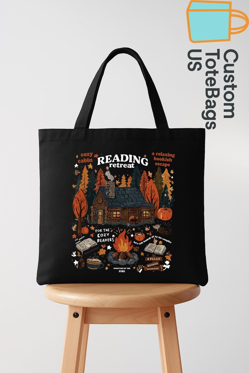 May include: Black tote bag featuring a cabin scene with a campfire and books. Text includes "Reading Retreat", "Cozy Cabin", and "For the Cozy Readers". The design is in autumnal colors.