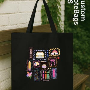 May include: Black tote bag featuring colourful illustrations of a school bus, rainbow, bow, and pencils. The bag has a rectangular shape with a handle. The text "BE KIND" is printed on the bag.