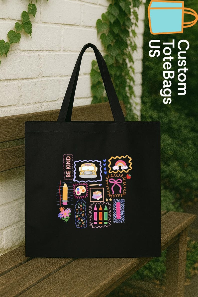 May include: Black tote bag featuring colourful illustrations of a school bus, rainbow, bow, and pencils. The bag has a rectangular shape with a handle. The text "BE KIND" is printed on the bag.