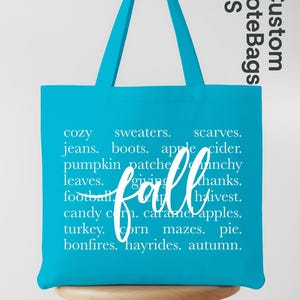 May include: A turquoise tote bag with white text listing fall-themed words like "cozy," "sweaters," and "pumpkin patch." The word "fall" is written in cursive. The bag is on a wooden stool.