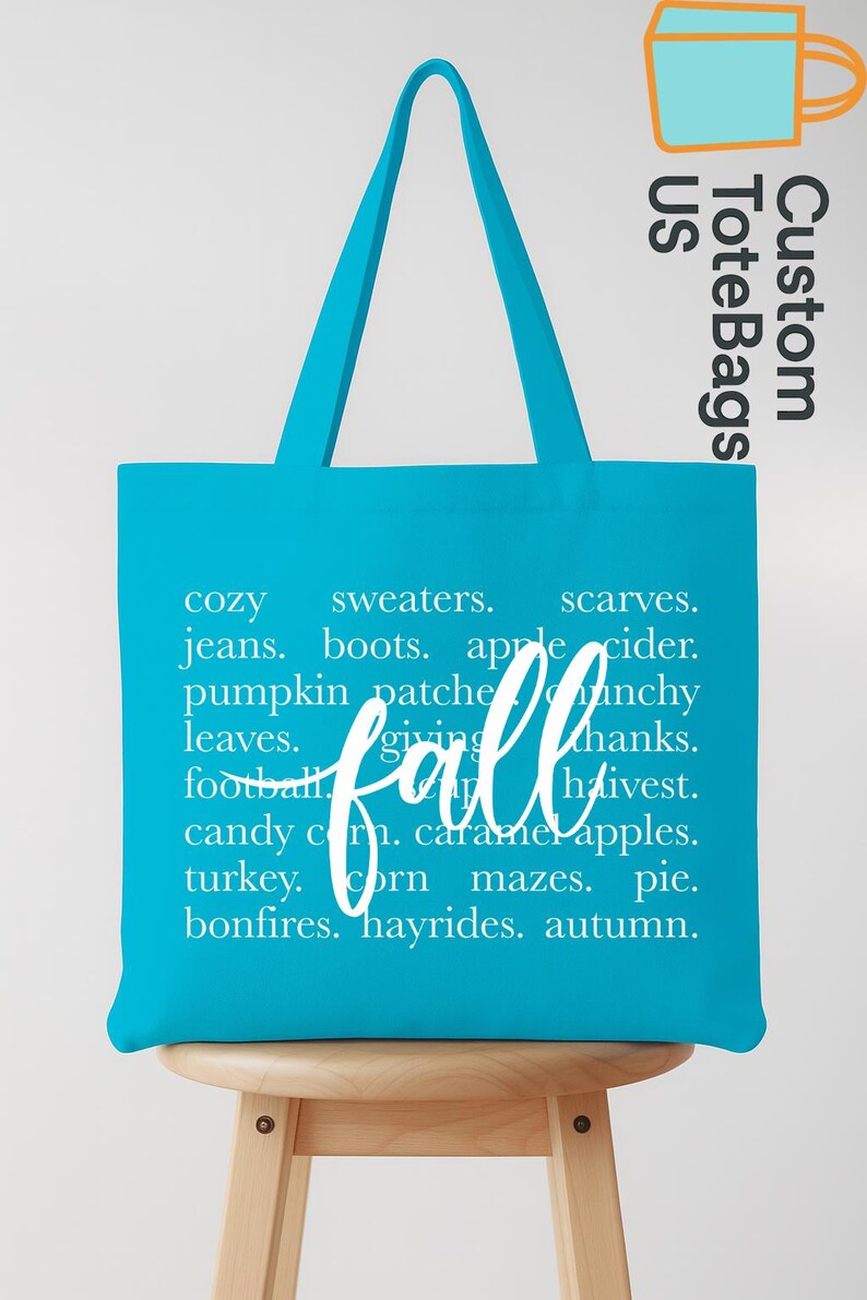 May include: A turquoise tote bag with white text listing fall-themed words like "cozy," "sweaters," and "pumpkin patch." The word "fall" is written in cursive. The bag is on a wooden stool.