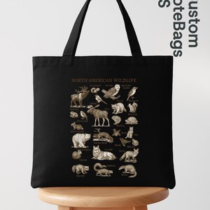 May include: Black tote bag with a detailed illustration of North American wildlife. The design includes various animals such as moose, owls, bears, and wolves, with each species labeled. The bag is displayed on a wooden stool.
