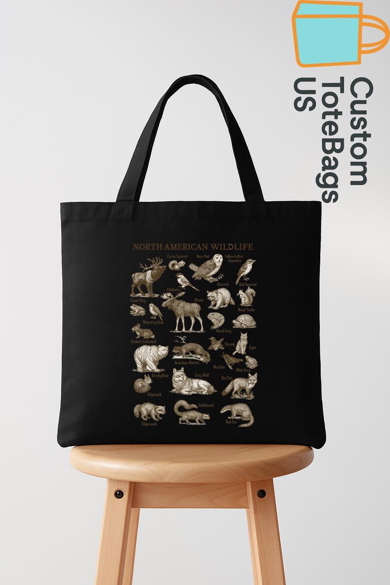 May include: Black tote bag with a detailed illustration of North American wildlife. The design includes various animals such as moose, owls, bears, and wolves, with each species labeled. The bag is displayed on a wooden stool.