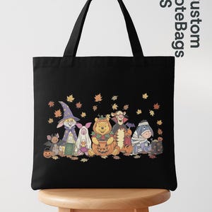 May include: A black tote bag featuring a graphic print of Winnie the Pooh characters dressed in Halloween costumes, including a wizard, ghost, pumpkin, vampire, and mummy, surrounded by falling autumn leaves on a black background.