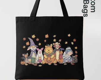 Peut inclure: Un sac fourre-tout noir avec une impression graphique des personnages de Winnie l'ourson déguisés pour Halloween, incluant un magicien, un fantôme, une citrouille, un vampire et une momie, entourés de feuilles d'automne sur fond noir.