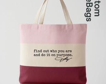 May include: A canvas tote bag featuring three horizontal color-blocked sections in light pink, cream, and deep burgundy. The center cream section displays the black text: find out who you are and do it on purpose. Dolly. It has two off-white handles.