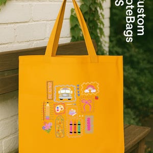 May include: A bright yellow tote bag featuring various colourful graphic designs, such as a school bus, rainbow, and pencils. The bag has long handles and the words "Custom ToteBags US" are printed on the side. The words "BE KIND" are also printed on the bag.