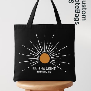 May include: Black tote bag with a white and orange sunburst design. The text "BE THE LIGHT" and "MATTHEW 5:14" are printed below the sun graphic. The bag has two black handles and is displayed on a wooden stool.
