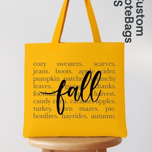 May include: A yellow tote bag with black text that reads "fall" in cursive and a list of fall-themed words. The bag has long handles and is sitting on a wooden stool. The words include "cozy", "sweaters", and "autumn."