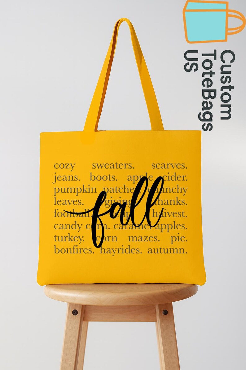 May include: A yellow tote bag with black text that reads "fall" in cursive and a list of fall-themed words. The bag has long handles and is sitting on a wooden stool. The words include "cozy", "sweaters", and "autumn."