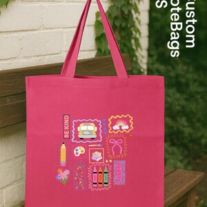 May include: A cerise tote bag featuring school-themed illustrations such as a bus, rainbow, and pencils. The bag has the text "BE KIND" and "Custom ToteBags US". The bag is displayed on a wooden bench. The bag is ideal for carrying books.