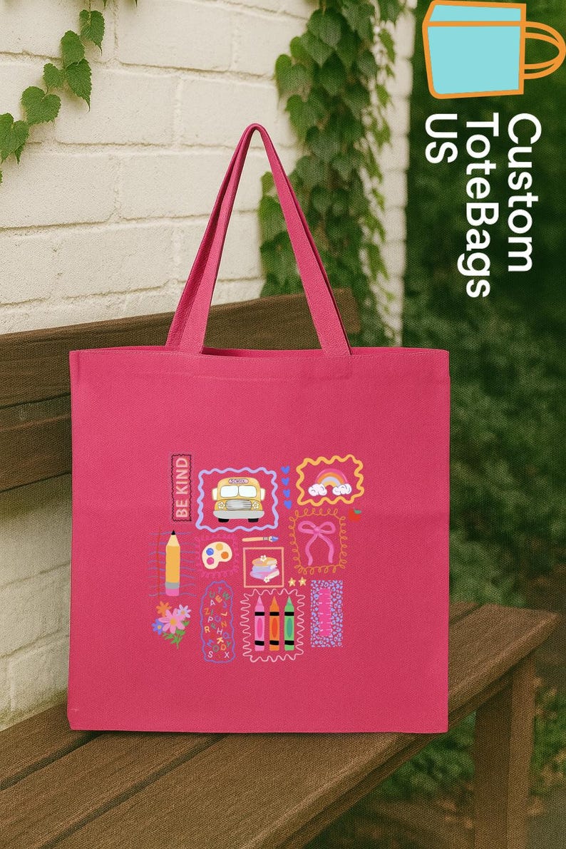 May include: A cerise tote bag featuring school-themed illustrations such as a bus, rainbow, and pencils. The bag has the text "BE KIND" and "Custom ToteBags US". The bag is displayed on a wooden bench. The bag is ideal for carrying books.