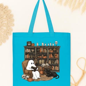 Puede incluir: Un bolso tote turquesa con una ilustraci&oacute;n de Halloween. Un fantasma lee en una silla frente a una estanter&iacute;a, con un gato negro, calabazas y calaveras. El bolso tiene asas turquesas.