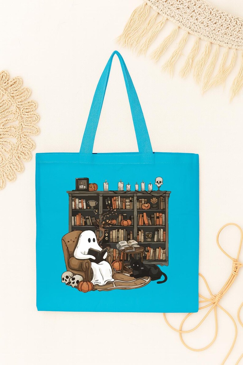 Puede incluir: Un bolso tote turquesa con una ilustraci&oacute;n de Halloween. Un fantasma lee en una silla frente a una estanter&iacute;a, con un gato negro, calabazas y calaveras. El bolso tiene asas turquesas.