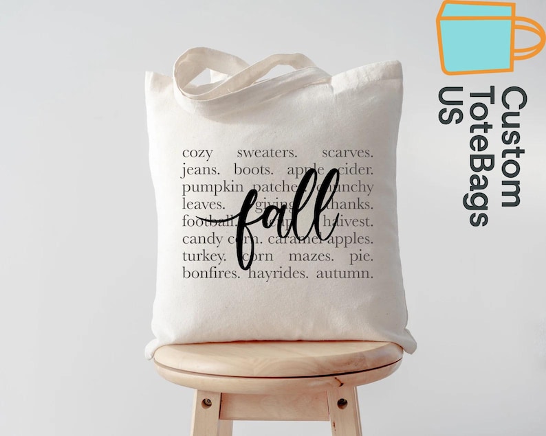 May include: A cream-colored tote bag with the word "fall" in a cursive font. The bag is printed with a list of fall-themed words like "sweaters," "pumpkin patch," and "hayrides." The bag is sitting on a wooden stool.