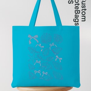 K&ouml;nnte beinhalten: Eine t&uuml;rkisblaue Tragetasche mit einem Muschel- und Schleifenmuster. Die Tasche hat eine rechteckige Form mit einem passenden t&uuml;rkisblauen Griff. Die Worte "Custom Tote Bags US" sind auf der Seite aufgedruckt. Die Tasche steht auf einem Holzschemel.