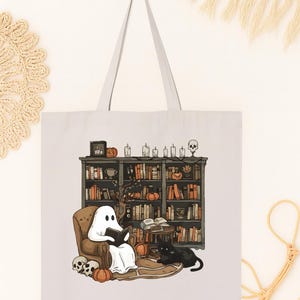 Puede incluir: Bolso tote blanco con una ilustraci&oacute;n de Halloween. Un fantasma lee en una silla frente a una estanter&iacute;a, con un gato negro, calabazas y calaveras. El bolso tiene un asa gris claro.