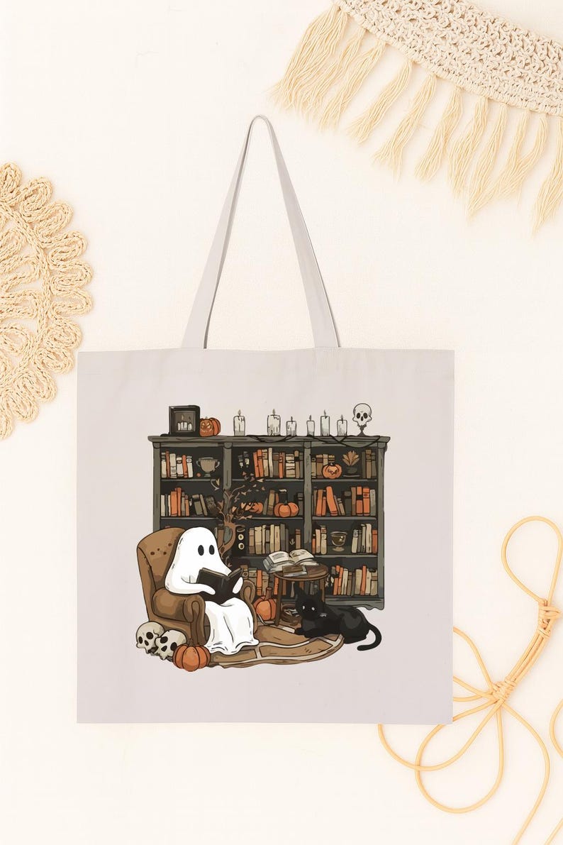 Puede incluir: Bolso tote blanco con una ilustraci&oacute;n de Halloween. Un fantasma lee en una silla frente a una estanter&iacute;a, con un gato negro, calabazas y calaveras. El bolso tiene un asa gris claro.