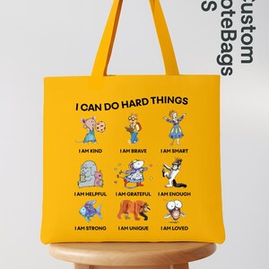 May include: Yellow tote bag with the text "I CAN DO HARD THINGS" and illustrations of cartoon animals with positive affirmations. The bag has a matching yellow handle and the words "Custom Tote Bags US" are printed on the side.