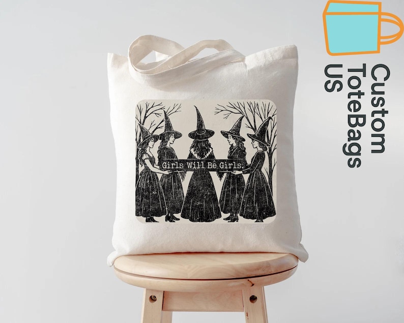 May include: A cream tote bag featuring a black and white illustration of five witches in long dresses and pointed hats, holding a banner with the text "Girls Will Be Girls." The bag has two handles and is placed on a wooden stool.