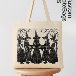 May include: A cream tote bag with a black and white illustration of five figures in witch hats and dresses, with the text "Girls Will Be Girls." The bag has long handles and a simple design.