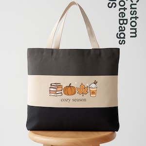 May include: A black, beige, and dark gray tote bag with cream-colored handles. The bag features a fall-themed graphic with a pumpkin, maple leaf, books, and a coffee cup. The words "cozy season" are printed below the graphic.