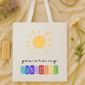 Peut inclure: Sac fourre-tout blanc avec un soleil jaune et le texte "you are my SUNSHINE" en lettres color&eacute;es. Le sac a des anses blanches et est pos&eacute; sur une surface beige. Un appareil photo et des fleurs sont en arri&egrave;re-plan.