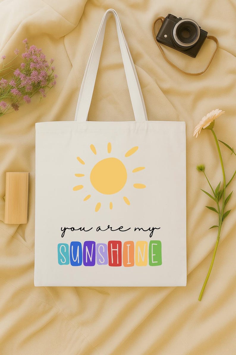 Peut inclure: Sac fourre-tout blanc avec un soleil jaune et le texte "you are my SUNSHINE" en lettres color&eacute;es. Le sac a des anses blanches et est pos&eacute; sur une surface beige. Un appareil photo et des fleurs sont en arri&egrave;re-plan.