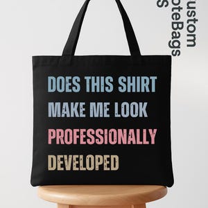 May include: A black tote bag with two shoulder straps featuring the text "DOES THIS SHIRT MAKE ME LOOK PROFESSIONALLY DEVELOPED" printed in light blue, pink, and yellow block letters. The bag is displayed on a light wooden stool.