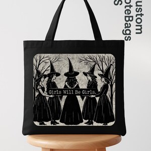 May include: Black tote bag with a graphic of five witches in black dresses and pointed hats, standing in a circle. The text "Girls Will Be Girls" is displayed below the witches. The bag has black handles.