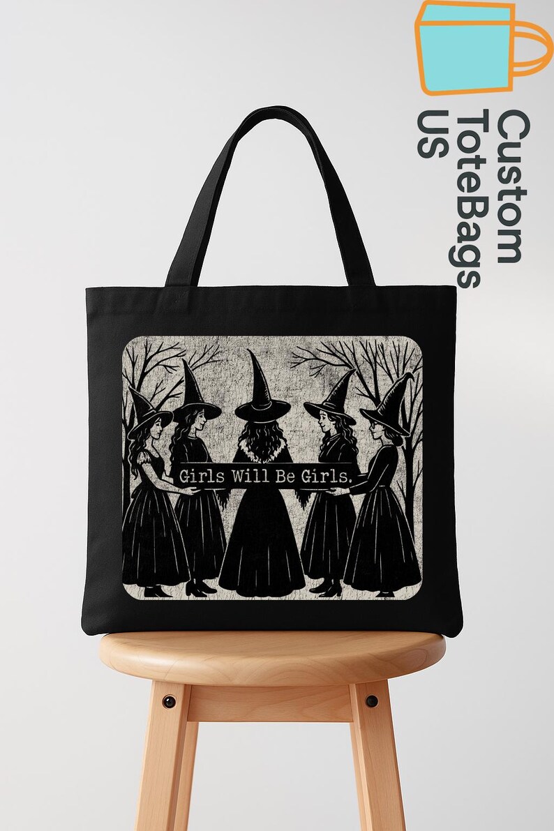 May include: Black tote bag with a graphic of five witches in black dresses and pointed hats, standing in a circle. The text "Girls Will Be Girls" is displayed below the witches. The bag has black handles.