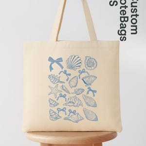 K&ouml;nnte beinhalten: Cremefarbene Tragetasche mit blauem Muschel- und Schleifenmuster. Die Tasche hat lange Henkel und wird auf einem Holzschemel pr&auml;sentiert. Der Text "Custom Tote Bags US" ist oben rechts zu sehen.