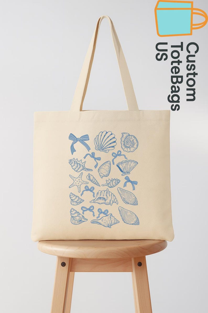 K&ouml;nnte beinhalten: Cremefarbene Tragetasche mit blauem Muschel- und Schleifenmuster. Die Tasche hat lange Henkel und wird auf einem Holzschemel pr&auml;sentiert. Der Text "Custom Tote Bags US" ist oben rechts zu sehen.