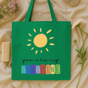 Peut inclure: Sac fourre-tout vert avec un soleil jaune et l'inscription "you are my sunshine". Le texte est en &eacute;criture noire, le mot "sunshine" est en lettres color&eacute;es. Le sac a de longues anses vertes.