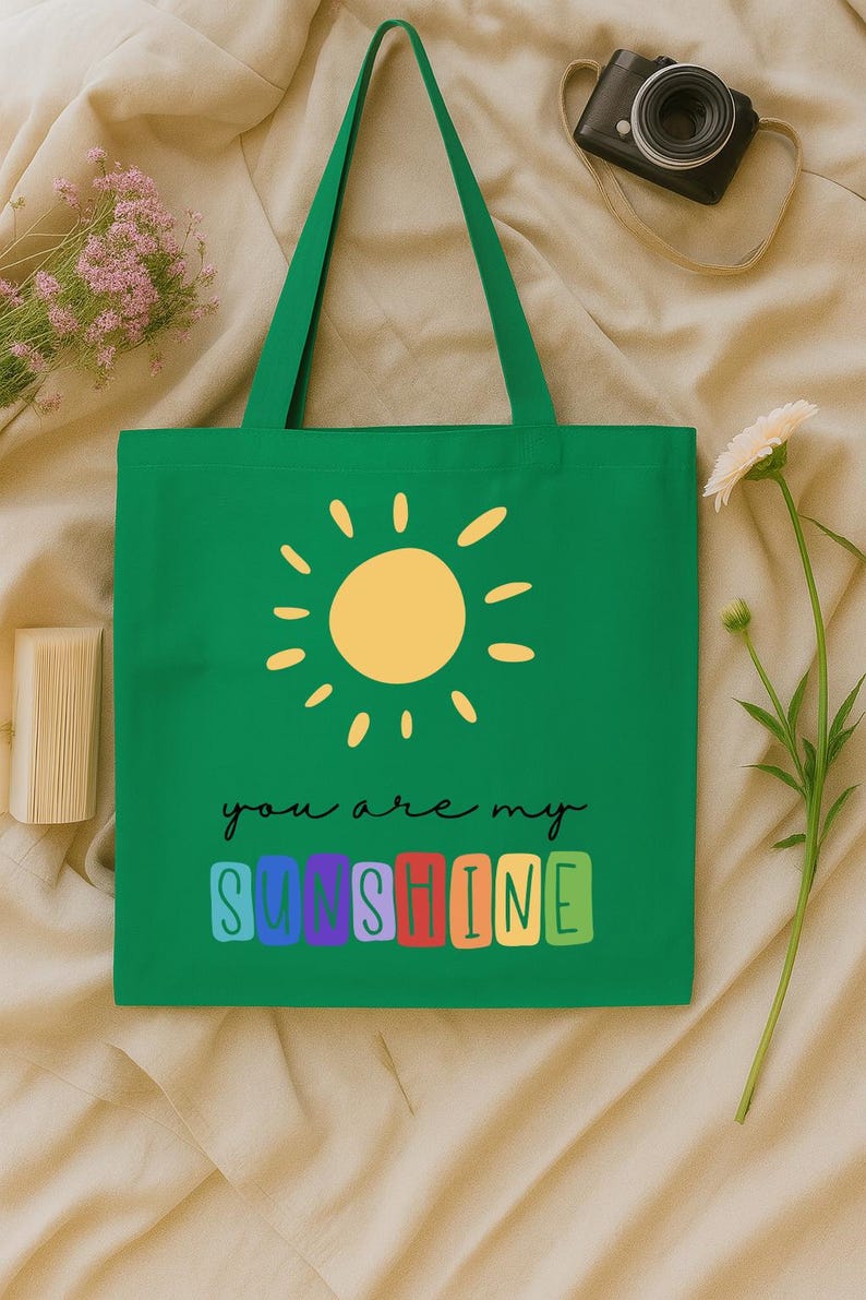 Peut inclure: Sac fourre-tout vert avec un soleil jaune et l'inscription "you are my sunshine". Le texte est en &eacute;criture noire, le mot "sunshine" est en lettres color&eacute;es. Le sac a de longues anses vertes.