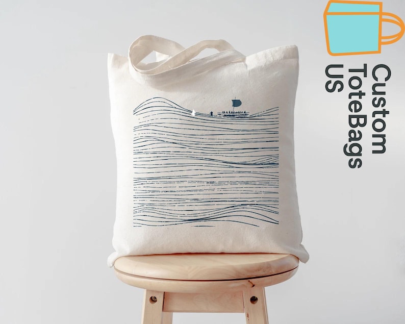 Puede incluir: Bolso tote de color crema con una ilustraci&oacute;n azul de un barco sobre l&iacute;neas onduladas. El bolso tiene un par de asas. El texto "Custom ToteBags US" est&aacute; impreso en el lateral.