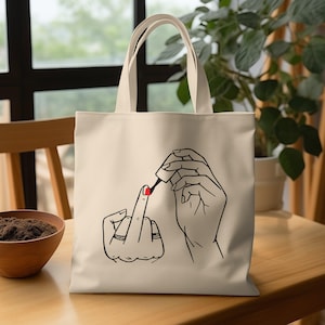 May include: Cream-colored tote bag featuring a black line drawing of hands. One hand is giving the middle finger, while the other hand is applying red nail polish to the middle finger. The bag has two cream-colored handles.