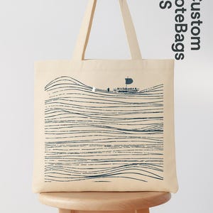 Puede incluir: Bolso tote de color crema con una ilustraci&oacute;n azul marino de un barco navegando sobre l&iacute;neas onduladas. El bolso tiene asas largas y el texto "Custom Tote Bags US" en orientaci&oacute;n vertical.