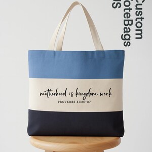 May include: A canvas tote bag with a three-tone design: light blue, cream, and navy. The bag features the text "motherhood is kingdom work" and "PROVERBS 31:25-27" in black script. The handles are a cream color.