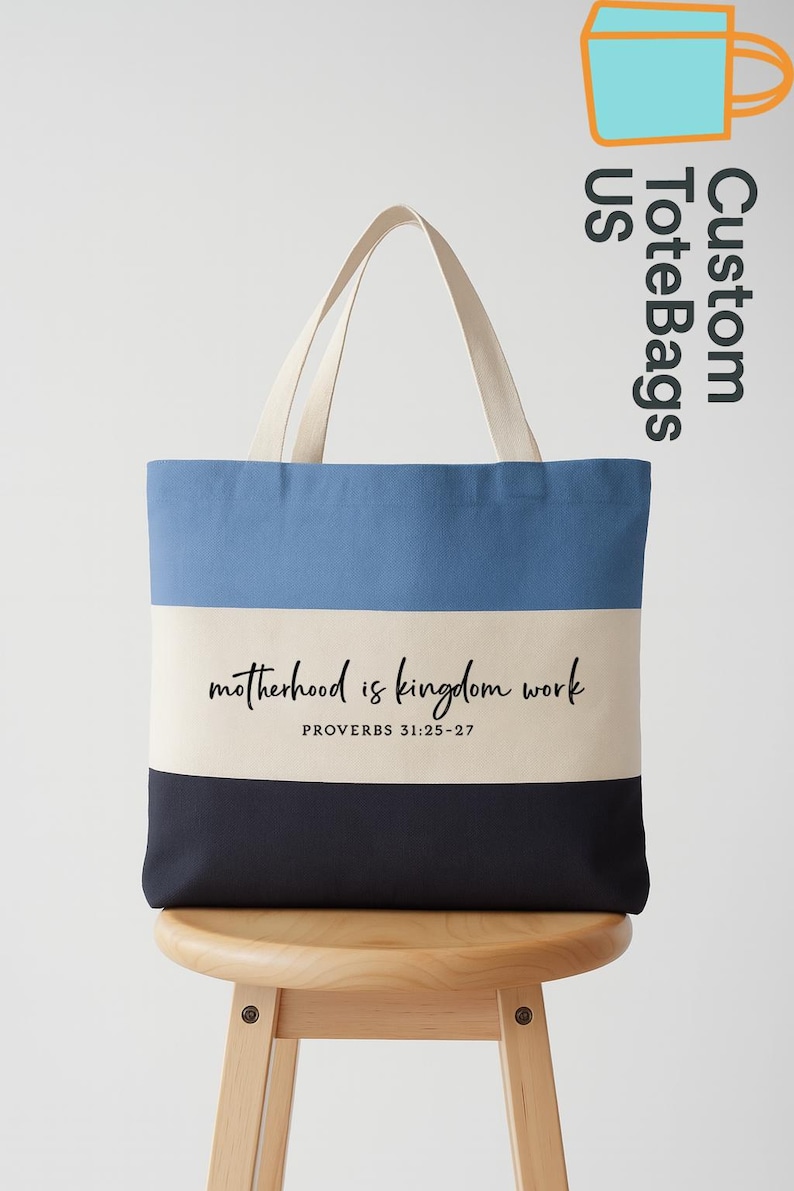 May include: A canvas tote bag with a three-tone design: light blue, cream, and navy. The bag features the text "motherhood is kingdom work" and "PROVERBS 31:25-27" in black script. The handles are a cream color.