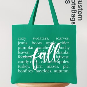 May include: A green tote bag with white text that reads "fall" in cursive. The bag lists fall-themed words like "cozy," "sweaters," "pumpkin patch," and "hayrides." The bag also has the text "Custom Tote Bags US".