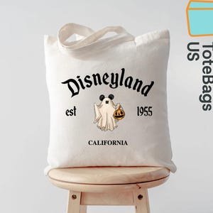 May include: A white canvas tote bag featuring the Disneyland logo with a ghost wearing Mickey Mouse ears and holding a jack-o'-lantern. The text "est 1955 CALIFORNIA" is also printed on the bag. The bag has a handle and is sitting on a wooden stool.