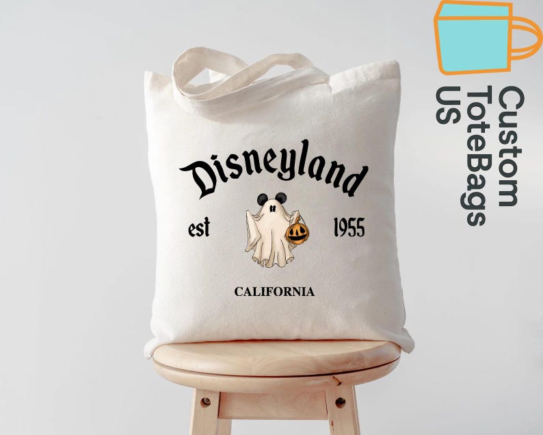 May include: A white canvas tote bag featuring the Disneyland logo with a ghost wearing Mickey Mouse ears and holding a jack-o'-lantern. The text "est 1955 CALIFORNIA" is also printed on the bag. The bag has a handle and is sitting on a wooden stool.