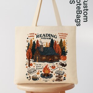 May include: Cream tote bag with a cabin scene and the words "READING retreat." The design includes a cabin, trees, a campfire, books, and the text "For the cozy readers."