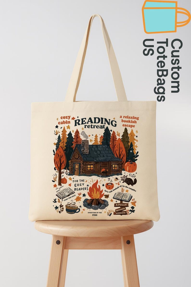 May include: Cream tote bag with a cabin scene and the words "READING retreat." The design includes a cabin, trees, a campfire, books, and the text "For the cozy readers."
